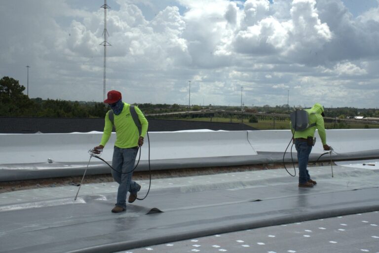 Two crew members spray-applying adhesive across a commercial flat roof